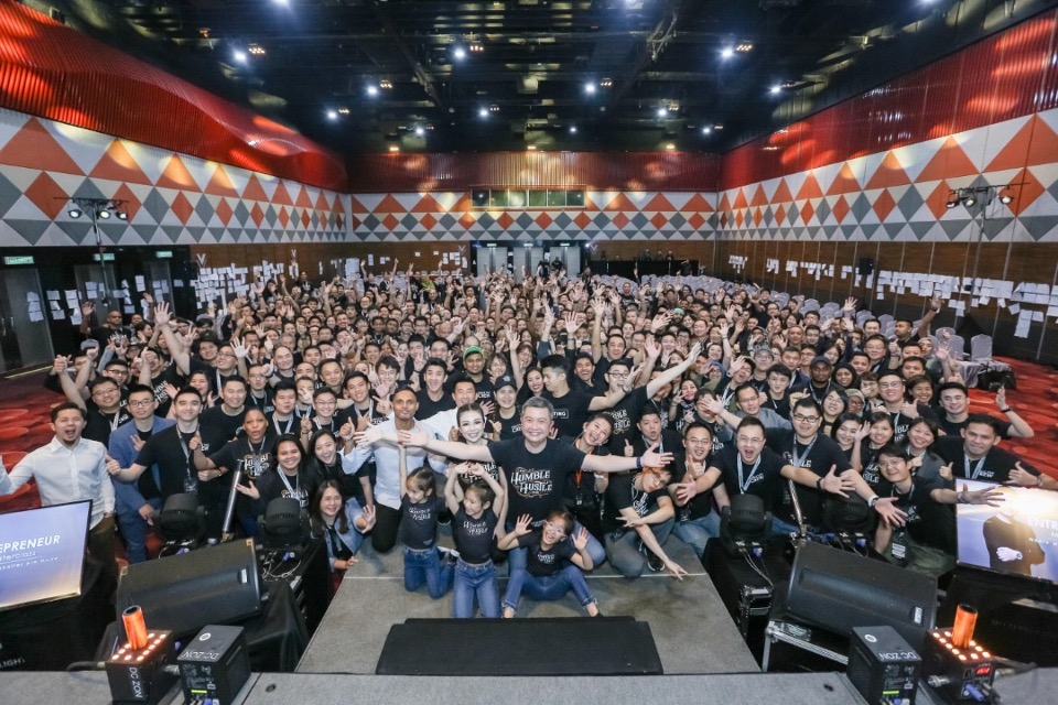 A packed theatrical hall of high-performers cheering with arms raised at a WORLDMASTERCLASS live event